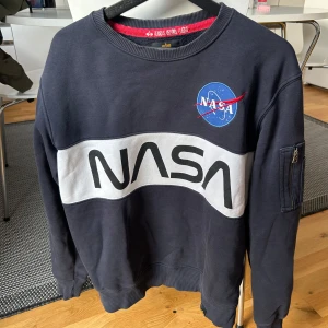 Mörkblå NASA sweatshirt från Alpha Industries - Säljer en snygg mörkblå sweatshirt från Alpha Industries med NASA-logga. Tröjan har en vit panel med stor NASA-text och en praktisk ficka med dragkedja på ärmen. Passar både män och kvinnor