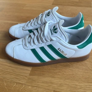 Vita och gröna Adidas Gazelle sneakers - Säljer ett par klassiska Adidas Gazelle sneakers i vitt med gröna detaljer och bruna sulor. Skorna har den ikoniska tre-randiga designen och snörning framtill. Perfekta för en sportig och stilren look.