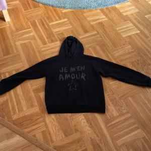 Svart hoodie från Gina Tricot - Säljer en svart hoodie från Gina Tricot med texten 'JE M'EN AMOUR' och en stjärna på framdiden💕Den är i fint skick💕Nypris kostar den 250 kr men jag säljer den för 100 kr💕Hör av er vid intresse💕💕