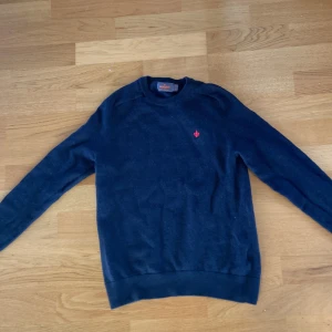 Mörkblå merinoull från Morris - Säljer en stilren mörkblå tröja från Morris i 100% merinoull. Tröjan har en liten röd broderad detalj på bröstet och långa ärmar. Perfekt för kyligare dagar. 