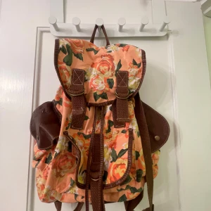 Blommig ryggsäck med bruna detaljer - Säljer en färgglad ryggsäck med blommigt mönster i orange och grönt. Väskan har bruna detaljer och justerbara axelremmar. Perfekt för att ge en färgklick till din outfit! Sliten på vänstra lilla facket. Knappen har lossnat på mitten facket. Tyvärr hål på baksidan vid axelremmen, men kan lagas. Köpt på Claire’s.