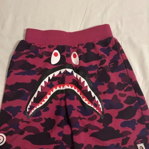 Kamouflagefärgade shorts från A Bathing Ape - Coola kamouflagefärgade shorts från A Bathing Ape i lila och rosa toner. De har ett unikt hajmotiv framtill och WGM-tryck på sidan. Perfekta för en streetwear-look med en bekväm passform. Använt typ 5 gånger så de är som helt nya. Dm för fler bilder!