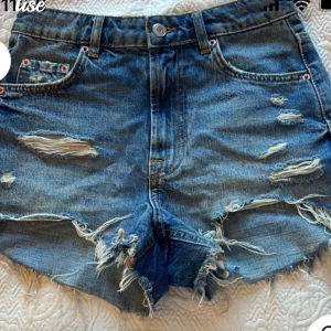 Blå jeansshorts med slitningar - Säljer ett par coola blå jeansshorts med slitningar och fransiga kanter. De har en klassisk femficksdesign och knappgylf. Perfekta för en avslappnad stil.