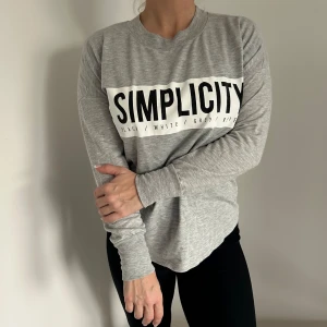 Grå långärmad tröja med text - Säljer en mysig grå långärmad tröja med texten 'SIMPLICITY' i svart på bröstet🩶