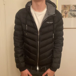 Svart dunjacka från Armani Exchange - Snygg svart dunjacka från Armani Exchange med huva och dragkedja. Jackan har en quiltad design och logotyp på bröstet. Perfekt för vintern/våren. Nypris 1800kr mitt pris 500kr. Inga defekter