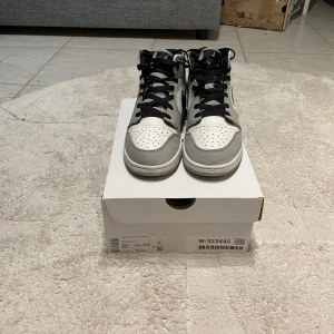 Nike Air Jordan 1 Mid i grå och vit - Snygga Nike Air Jordan 1 Mid sneakers i en stilren kombination av grått och vitt. Skorna har klassisk snörning och den ikoniska Nike-loggan i svart på sidan. Perfekta för dig som vill ha en sportig och trendig look.
