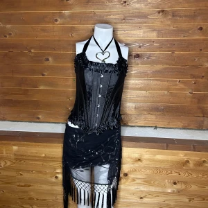 Svart korsett med volangdetaljer - Snygg svart korsett med volangdetaljer och knäppning framtill. Den har en halterneck-design och är tillverkad i ett glansigt material som ger en lyxig känsla. Perfekt för att skapa en dramatisk look.