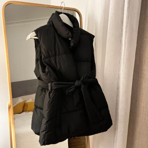 Svart väst med bälte - Snygg svart väst med hög krage och knytbälte i midjan. Perfekt för kyliga dagar när du vill hålla stilen. Västen har en design som ger extra värme och komfort.