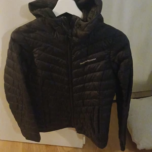 Svart dunjacka från Peak Performance - Säljer en svart dunjacka från Peak Performance. Jackan har en huva och dragkedja framtill. Perfekt för kyliga dagar med sin quiltade design som håller värmen. En stilren och funktionell jacka för alla tillfällen.