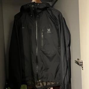 Svart regnjacka från Haglöfs - Säljer en svart regnjacka från Haglöfs med huva och dragkedja framtill. Jackan har en bröstficka med dragkedja och är perfekt för regniga dagar. Den är designad för att hålla dig torr och bekväm.