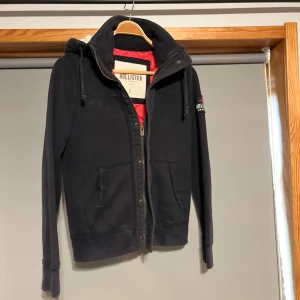 Fodrad cardigan jacka - Säljer en mörkblå cardigan jacka från Hollister med dragkedja och broderad logga på ärmen. Den har en mysig fodrad huva och långa ärmar. Perfekt för kyliga dagar!