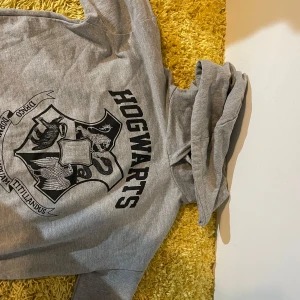 Grå hoodie med Hogwarts-tryck - Säljer en grå hoodie med stort Hogwarts-tryck på framsidan. Perfekt för alla Harry Potter-fans! Hoodien har en bekväm passform och är tillverkad i mjukt material. Perfekt för mysiga dagar hemma eller som en cool vardagströja.