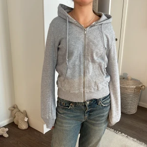 Grå hoodie med dragkedja - Säljer en stilren grå hoodie med dragkedja och huva. Perfekt för en avslappnad look. Tröjan har långa ärmar och ribbade muddar vid ärmslut och nederkant. Passar bra till jeans eller mjukisbyxor.
