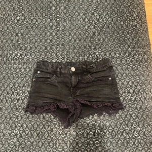 Svarta jeansshorts med spetsdetaljer - Snygga svarta jeansshorts med spetsdetaljer längst ner. Perfekta för en cool och avslappnad stil. Shortsen har fickor både fram och bak samt en knapp och dragkedja framtill.