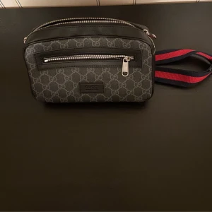 Gucci beltbag - Snygg svart axelväska från Gucci med klassiskt monogrammönster. Väskan har en justerbar axelrem i rött och svart samt en praktisk dragkedja framtill. Perfekt för att bära med sig det viktigaste med stil.