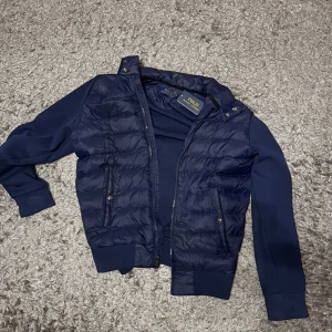 Blå jacka från Polo Ralph Lauren - Snygg blå jacka från Polo Ralph Lauren med quiltad framsida och dragkedja. Jackan har långa ärmar och ribbade muddar. Perfekt för en stilren look.