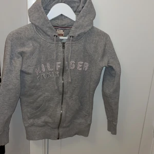 Grå hoodie från Hilfiger Denim - Grå hoodie från Hilfiger Denim med zipper och luva. Tröjan har ett tryck med texten 'Hilfiger Denim' på framsidan och praktiska fickor. 