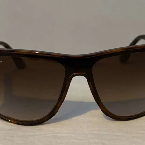 Bruna solglasögon från Ray-Ban - Snygga solglasögon från Ray-Ban med en klassisk brun båge. De har en stilren design med Ray-Ban-loggan på skalmarna. Perfekta för soliga dagar och kommer med ett svart fodral, putsduk, original box.