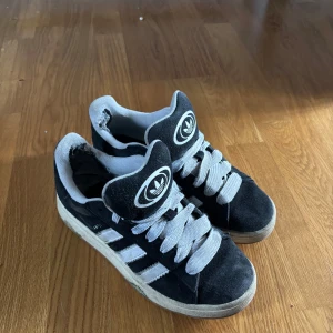 Adidas campus 00 - Säljer ett par svarta sneakers från Adidas med vita detaljer och snörning. Skorna har en klassisk design med tre vita ränder på sidan och en vit sula. Perfekta för en avslappnad stil. 