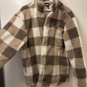Rutig teddyjacka från H&M - Mysig teddyjacka från H&M i brunt och beige rutmönster. Jackan har långa ärmar och en praktisk bröstficka. Perfekt för kyliga dagar med sin mjuka och varma känsla.