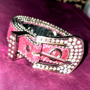 Glittrigt rosa bälte med nitar - Snyggt rosa bälte med glittriga detaljer och nitar. Perfekt för att ge din outfit en extra touch. Spännet är dekorerat med gnistrande stenar som verkligen fångar ögat.