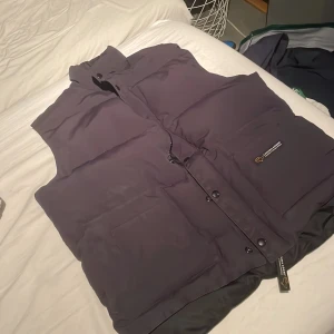 Svart dunväst från Canada Goose - Säljer en svart dunväst från Canada Goose. Västen har en dragkedja och knappar framtill samt två stora fickor. Perfekt för kyligare dagar.