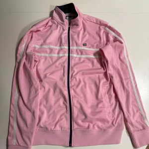 Weekend Offender - Säljer en snygg rosa jacka från Weekend Offender med vita ränder längs ärmarna. Jackan har dragkedja framtill och en sportig look. Perfekt för en avslappnad stil.
