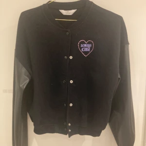 Svart lätt jacka från H&M med hjärtdetaljer - Säljer en svart tunn jacka från H&M med knappar framtill och texten 'ALWAYS CARE' i ett hjärta på bröstet och ryggen. Ärmarna har en kontrasterande design. Jackan är klar svart i färgen (ljuset på bilden gör färgen gråsvart men den är svart)