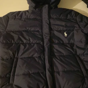 Svart pufferjacka från Ralph Lauren - Snygg svart pufferjacka från Ralph Lauren med dragkedja och broderad logga på bröstet. Jackan har långa ärmar och är perfekt för kyligare väder.