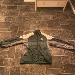 Green Bay Packers vindjacka från  Reebok  - Säljer en grön och vit Green Bay Packers vindjacka. . Jackan har en dragkedja framtill och långa ärmar. Den har en Packers logga på bröstet och en röd detalj på ärmen. Perfekt för höst och vår. Kom med bud!