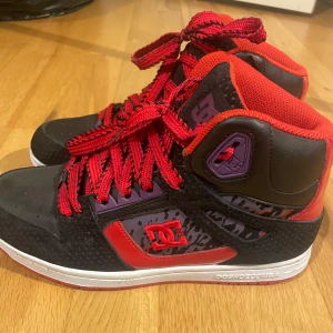 Svarta och röda sneakers från DC - Snygga höga sneakers från DC i svart och rött med coola detaljer. Skorna har en robust design med snörning och en vit sula. Perfekta för en streetwear-look.
