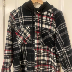 Rutig jacka med huva från Zara - Säljer en cool rutig jacka från Zara med svart huva. Jackan har en avslappnad passform och är perfekt för en casual look. Den har långa ärmar och en klassisk röd, svart och vit färgkombination.