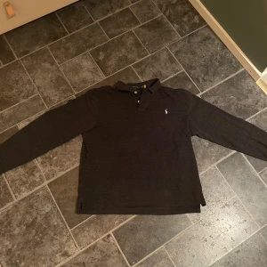 Grå pikétröja från Ralph Lauren - Säljer en stilren grå pikétröja från Ralph Lauren med klassisk krage och knappar. Tröjan har en broderad logotyp på bröstet och är långärmad. Perfekt för en avslappnad men ändå elegant look. Står L i storlek men skulle säga mer av en M Bra skick, inga flaws eller stains.