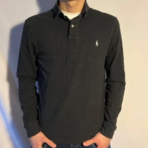 Svart långärmad pikétröja från Ralph Lauren - Säljer en stilren svart långärmad pikétröja från Ralph Lauren i slim fit. Tröjan har en klassisk krage och knappar framtill, med det ikoniska logotypbroderiet på bröstet. Perfekt för en elegant och avslappnad look.