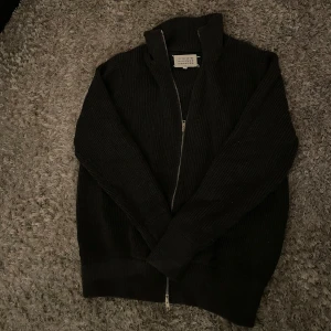 Fullzip från Maison Margiela - Snygg svart ribbad tröja från Maison Margiela med dragkedja och hög krage. Perfekt för kyligare dagar och ger en stilren look. Passar bra till jeans eller chinos.