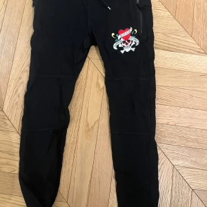 Svarta byxor från Ed Hardy - Snygga svarta byxor från Ed Hardy med ett coolt broderat motiv av en dödskalle och hjärta på sidan. Byxorna har dragkedja och snörning i midjan för en bekväm passform.