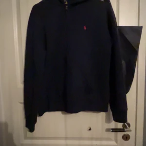 Marinblå hoodie från Ralph Lauren - Säljer en stilren marinblå hoodie från Ralph Lauren med dragkedja och det klassiska röda logotypbroderiet på bröstet. Perfekt för en avslappnad look.