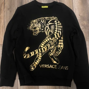 Svart tröja från Versace Jeans med tigertryck - Snygg svart tröja från Versace Jeans med ett iögonfallande guldtryck av en tiger på framsidan. Tröjan har långa ärmar och en klassisk rund halsringning. Perfekt för dig som vill sticka ut med stil.