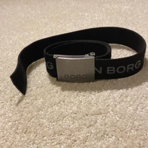 Svart bälte från Björn Borg - Snyggt svart bälte från Björn Borg med en stilren silverfärgad spänne. Bältet har Björn Borg-loggan tryckt längs hela remmen. Perfekt för att ge en sportig touch till din outfit.