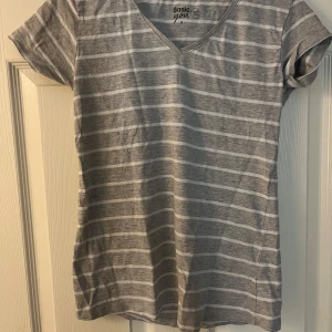 Grå och vit randig t-shirt från Basic You - Säljer en grå och vit randig t-shirt från Basic You i ekologisk bomull. T-shirten har en v-ringad hals och korta ärmar, perfekt för en avslappnad stil.