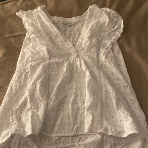 Vit blus från Rosebud - Säljer en söt vit blus från Rosebud med v-ringning och volangdetaljer. Blusen har ett broderat mönster och är ärmlös, perfekt för varmare dagar. Köpt från second hand för 90kr. Väldigt skön och ha speciellt på sommaren/sensommaren.