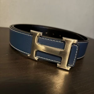 Blått skärp med H-spänne - Snyggt blått skärp med ett stort H-format spänne i silver. Skärpet är tillverkat i skinn och har en stilren design med vita sömmar längs kanterna. Perfekt för att ge en elegant touch till din outfit.