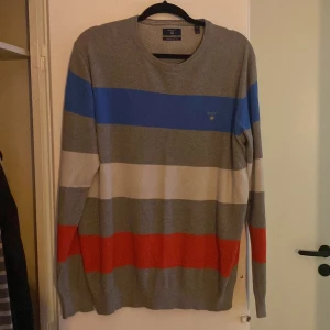 Randig tröja från Gant - Säljer en snygg randig tröja från Gant i premium bomull. Tröjan har breda ränder i grått, blått, vitt och rött. Den är långärmad och har en normal passform. Perfekt för en avslappnad stil.