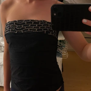 Svart axelbandslös klänning med pärlor - Elegant svart axelbandslös klänning med dekorativa pärlor längs överdelen. Perfekt för en kväll ute eller en speciell tillställning. Klänningen har en tight passform som framhäver figuren.