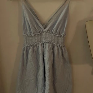 Ljusblå klänning med smockdetaljer - Charmig ljusblå klänning med smockdetaljer i midjan och v-ringad hals. Klänningen har tunna axelband och en luftig passform, perfekt för varma dagar.