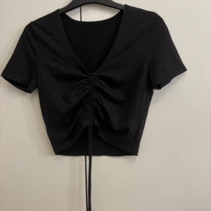 Svart kortärmad topp med knytband - Snygg svart kortärmad topp med v-ringning och knytning framtill. Aldrig använd bara legat o dammat i garderoben går att använda roll