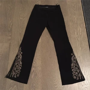 lågmidjade bootcut - Snygga svarta byxor från Miss Etam med vackra broderade blommor i guld längs benen. Perfekta för en festlig kväll eller när du vill känna dig extra fin. Byxorna har en elegant lågmidjade bootcut-stil mocca tyg som ger en smickrande siluett. De är i mycket bra skick och redo att bäras till nästa event! Midjemått ca 36cm innerbenslängd 83cm