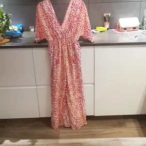 Leopardmönstrad långklänning med v-ringning - Byxklänning med leopardmönster i rött och vitt. Klänningen har en djup v-ringning och korta ärmar, perfekt för en somrig look. Den är tillverkad i ett lätt och luftigt material som ger en bekväm passform.