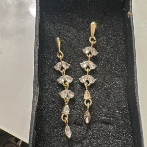 Guldörhängen med glittriga stenar - Eleganta guldörhängen med flera glittriga stenar i en hängande design. Perfekta för att ge en touch av glamour till din outfit. De har en smidig stängning och en vacker form som fångar ljuset fint. En sten saknas på det högra örhänget därav priset🥰
