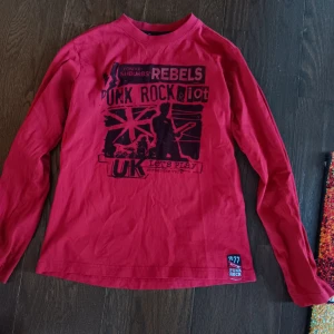 Röd långärmad tröja med punkmotiv - Säljer en cool röd långärmad tröja med tryck av punkmotiv. Är oxå mörkare röd än vad som syns på bilerna. Storlek ca S. Säljer pga passar ej mig som jag skulle önska. Tröjan har en rundad hals och är i en avslappnad passform.